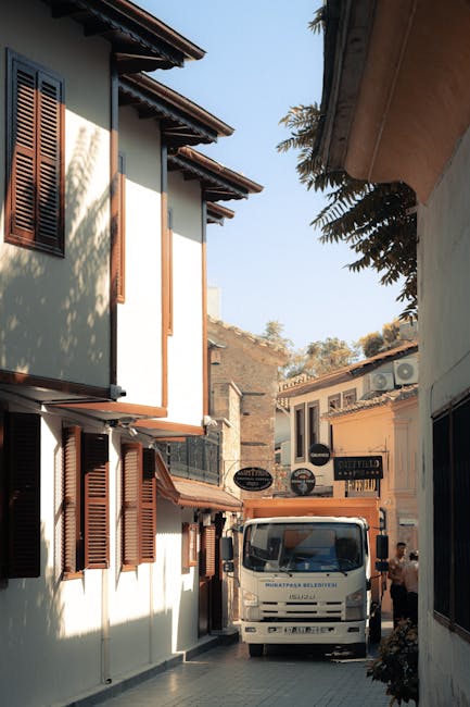A narrow alleyway between two multi-storey buildings with white and beige facades, featuring wooden window shutters and balconies. A white delivery van with visible branding is parked at the end of the alley, partially blocking the passage. Several cardboard boxes and packing materials are placed nearby, indicating a home relocation or moving process. A person can be seen near the van, possibly involved in loading or unloading furniture and boxes. The scene is lit by natural daylight, with some foliage visible overhanging from the right side. This image exemplifies a professional moving or removal service, such as those offered by Man and Van Kennington, coordinating transport logistics in residential areas.