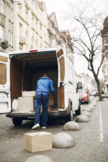A man wearing a blue jacket and dark pants is loading large white furniture pieces into the back of a white commercial van parked on a city street. The van's rear doors are open, revealing the interior lined with wooden panels, and the man is carefully placing the items inside. There is a cardboard box on the cobblestone pavement nearby, which is dotted with rounded stone bollards. In the background, multi-story residential buildings with ornate facades and balconies line the street, and a leafless tree stands visible against an overcast sky. This scene depicts home relocation or furniture transport as part of a professional removal service, with the man and Van Kennington handling packing and moving tasks efficiently and securely.