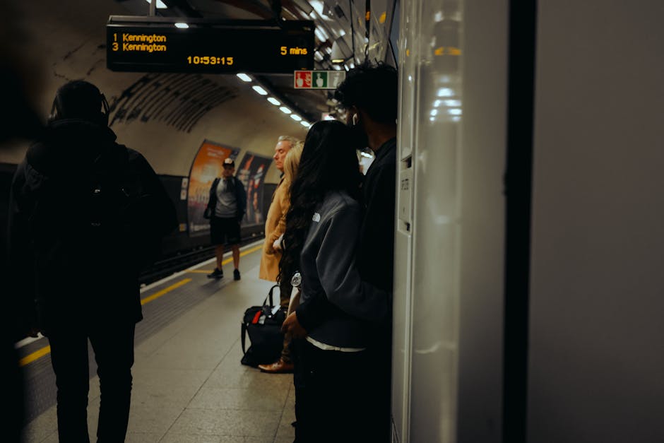 Photograph of a busy underground train station platform during daytime, with several passengers waiting for a train. Some individuals are standing close to the platform edge, while others are further back, holding luggage or backpacks. A digital display overhead shows upcoming train destinations, including Kennington, with scheduled arrival times. The station interior features curved ceiling panels with embedded lighting, and the edge of a train vehicle is visible on the right side of the image. The lighting is artificial, casting a slight glow on the commuters and the surrounding environment. This scene illustrates aspects of urban travel and busy transit environments, relevant to home relocation logistics where planning and timing are crucial, as seen in moving services provided by companies like Man and Van Kennington. The image captures the process of waiting for transport, akin to arranging furniture transport or packing and moving during house removals in the Kennington area. Such details underscore the importance of smooth loading, travel, and unloading processes involved in moving logistics.