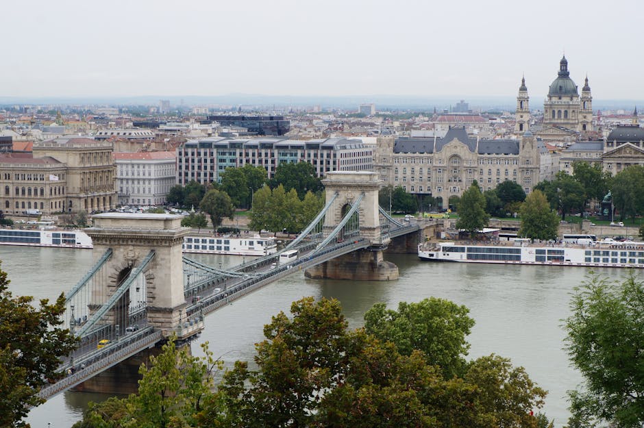 A panoramic view of a cityscape featuring the iconic Chain Bridge crossing the River Danube in Budapest, Hungary. The bridge has large stone towers with decorative architectural details and suspension