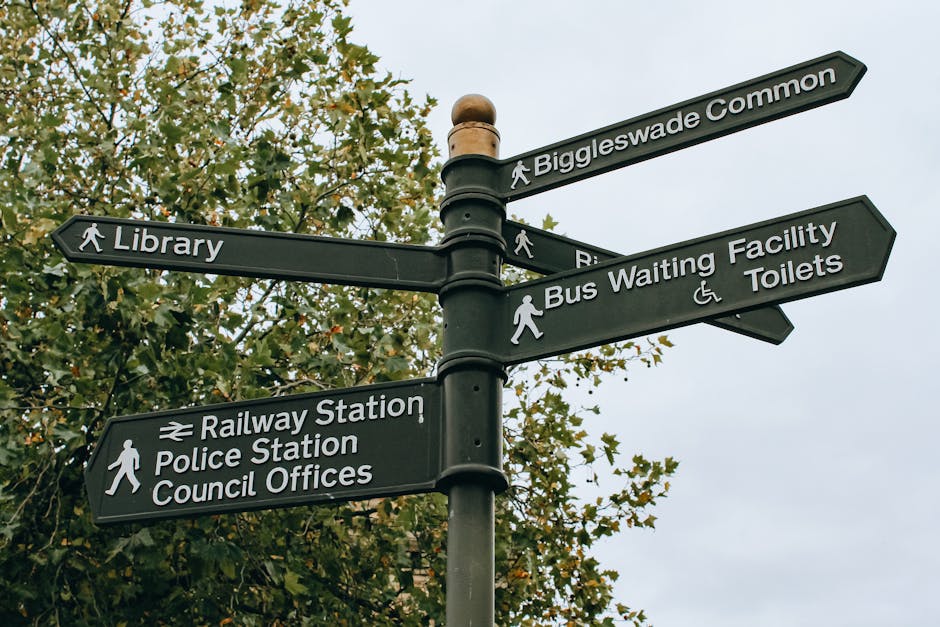 A black metal signpost with multiple directional signs is situated outdoors against a background of green leafy trees and a cloudy sky. The signs indicate directions to various local amenities, including 'Biggleswade Common', 'Library', 'Bus Waiting Facility and Toilets', 'Railway Station', 'Police Station', and 'Council Offices'. Each sign features white text and icons to denote pedestrian pathways, with some signs also showing symbols for accessible facilities. The signpost is positioned on a paved or paved-like surface, often used in residential or town centre areas for navigation. This scene exemplifies typical urban signage used to guide residents and visitors within a town or community, supporting efficient movement and local service access, which is relevant to home relocation and moving services like those offered by Man and Van Kennington, especially when planning for large item removals or navigating to designated collection points.