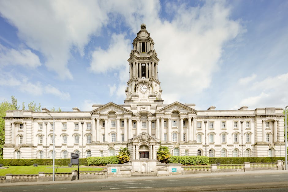 Exterior view of a historic, multi-story government building with classical architectural features, including a central clock tower with a spire, multiple arched windows, and ornate stone detailing. The building is constructed from light-colored stone and is surrounded by green bushes and small trees. The street in front has a paved road, and there are pedestrian features such as street signs and a fence. The sky above is mostly clear with some scattered clouds, and daylight illuminates the facade. The image showcases a setting suitable for house removals or home relocation services, emphasizing the importance of proper planning and permits when moving bulky items from such a building, as guided by Lambeth Council rules for Kennington bulky item removals. Man and Van Kennington may assist with furniture transport and packing and moving logistics in this area.