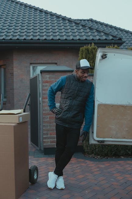 A man dressed in a black and blue jacket, black trousers, white sneakers, and wearing a grey cap stands outside a residential property during daylight. He is positioned next to an open white moving van with a beige panel on its side, which is parked on a paved driveway in front of a brick house with a dark tiled roof. In the foreground, there are cardboard boxes, some on a small hand truck, indicating a packing and loading process associated with home relocation or furniture transport. The man appears to be observing or preparing to assist with the loading or unloading of items as part of the house removals services carried out by Man and Van Kennington. The scene is well-lit with natural daylight, and the background includes a door and window of the house, alongside some greenery, suggesting a typical suburban setting for house moves in the Kennington area.