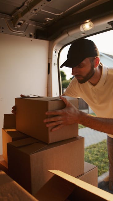 A man wearing a black cap and a white t-shirt is inside a van, carefully stacking and arranging several cardboard boxes during a home relocation process. The boxes vary in size and are organized on the floor of the vehicle, with some placed on top of others. The van's interior is illuminated by natural light coming through an open rear door, revealing a grassy outdoor area beyond. The man is focused on ensuring the boxes are securely loaded, representing the packing and loading stage of furniture transport and house removals. This scene, captured by Man and Van Kennington, illustrates professional moving services involved in packing and logistics for a residential move in Kennington as seen on the webpage https://manandvankennington.com.
