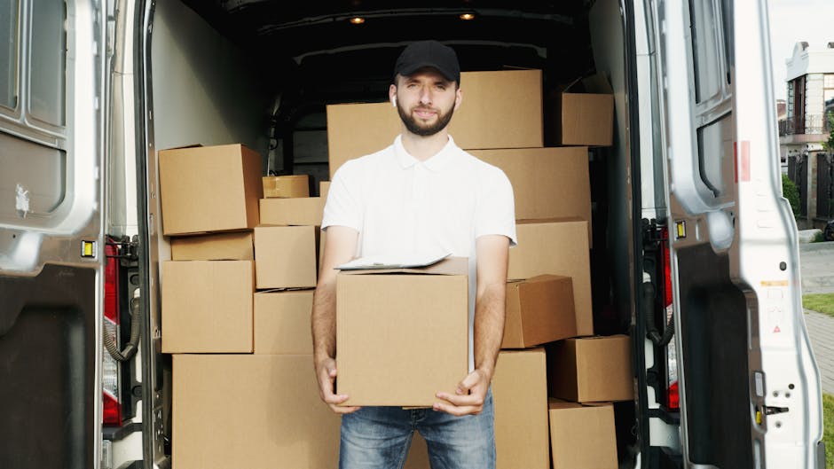 A man dressed in a black and blue jacket, black trousers, white sneakers, and wearing a grey cap stands outside a residential property during daylight. He is positioned next to an open white moving van with a beige panel on its side, which is parked on a paved driveway in front of a brick house with a dark tiled roof. In the foreground, there are cardboard boxes, some on a small hand truck, indicating a packing and loading process associated with home relocation or furniture transport. The man appears to be observing or preparing to assist with the loading or unloading of items as part of the house removals services carried out by Man and Van Kennington. The scene is well-lit with natural daylight, and the background includes a door and window of the house, alongside some greenery, suggesting a typical suburban setting for house moves in the Kennington area.