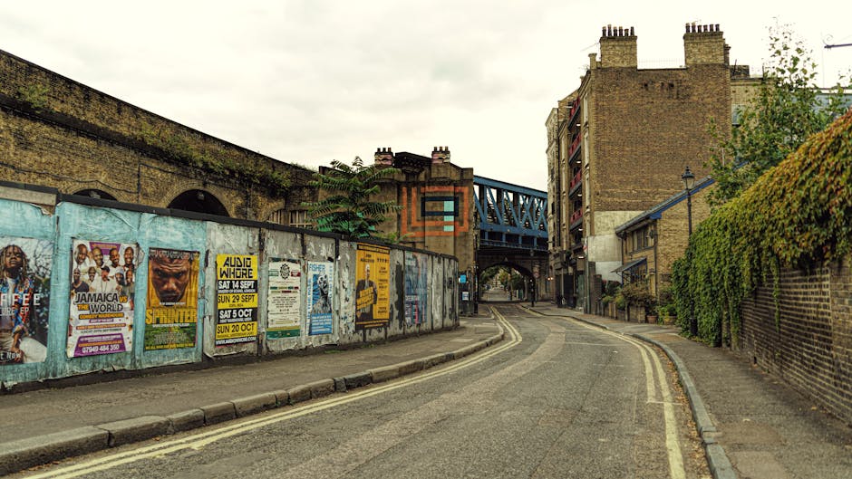 A narrow street in an urban area during daytime, with a lightly curved asphalt road featuring double yellow lines along its edge. On the left side, a weathered metal fence covered in various posters and advertisements separates the pavement from a brick wall, with posters promoting events and music gigs visible. Behind the fence, part of a brick building and an overpass with a blue metal bridge are apparent, indicating a transport or railway structure above the street. On the right, a tall brick wall is adorned with green foliage, and a narrow pavement runs alongside it. The scene exhibits overcast lighting with a cloudy sky, typical of a typical city environment suitable for house removals and relocation logistics, supporting the visual context of furniture transport and moving services like those offered by Man and Van Kennington.
