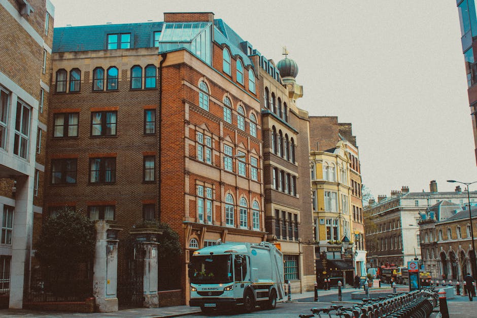 A city street scene showing a Man and Van Kennington vehicle parked on the pavement near a brick and stone building with multiple large, arched windows. The white van is positioned close to the entrance of a property, suggesting a home relocation or furniture transport process. Several cardboard boxes, wrapped furniture, and packing materials are visible on the pavement, indicating ongoing packing or loading activities. The surroundings include other historic and modern buildings, a lamppost, and bike racks along the sidewalk under natural daylight, with some vehicles and pedestrians in the background. The scene captures an urban environment typical for professional removals and moving services, with visible equipment such as dollies and straps possibly being used for safe loading and transportation in coordination with [COMPANY_NAME].