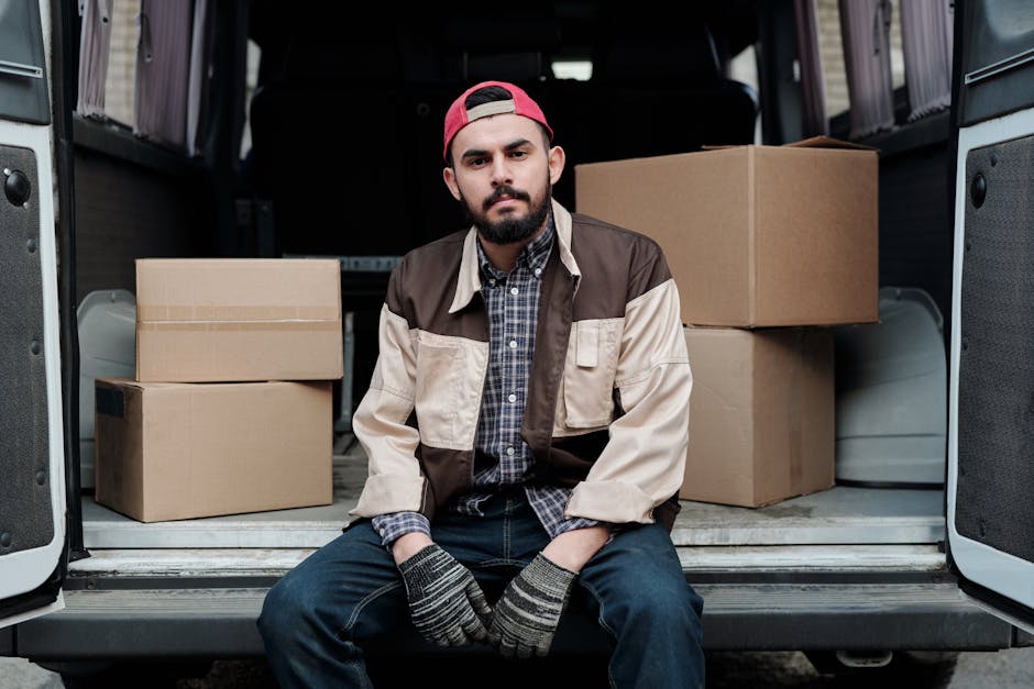 A young man with a beard, wearing a red cap, a beige and brown jacket over a checked shirt, and dark jeans, sits on the open back of a white van used for home relocation. Inside the van, there are several cardboard boxes in various sizes, some stacked on top of each other, with clear packing tape visible on their edges. The boxes are placed on the van’s interior floor, which is partly visible along with the vehicle’s rear doors. The man appears to be resting or pausing during the furniture transport and packing process, which is part of a professional removals service by Man and Van Kennington. The scene is well-lit, with natural light highlighting the arrangements of boxes and the man's neutral expression, reflecting the logistical aspect of loading for a house move or furniture transport. The proximity to the open vehicle emphasizes the loading process involved in home relocation services, with the focus on packing materials, the carrying of goods, and the logistical planning typical of removals undertaken by [COMPANY_NAME].