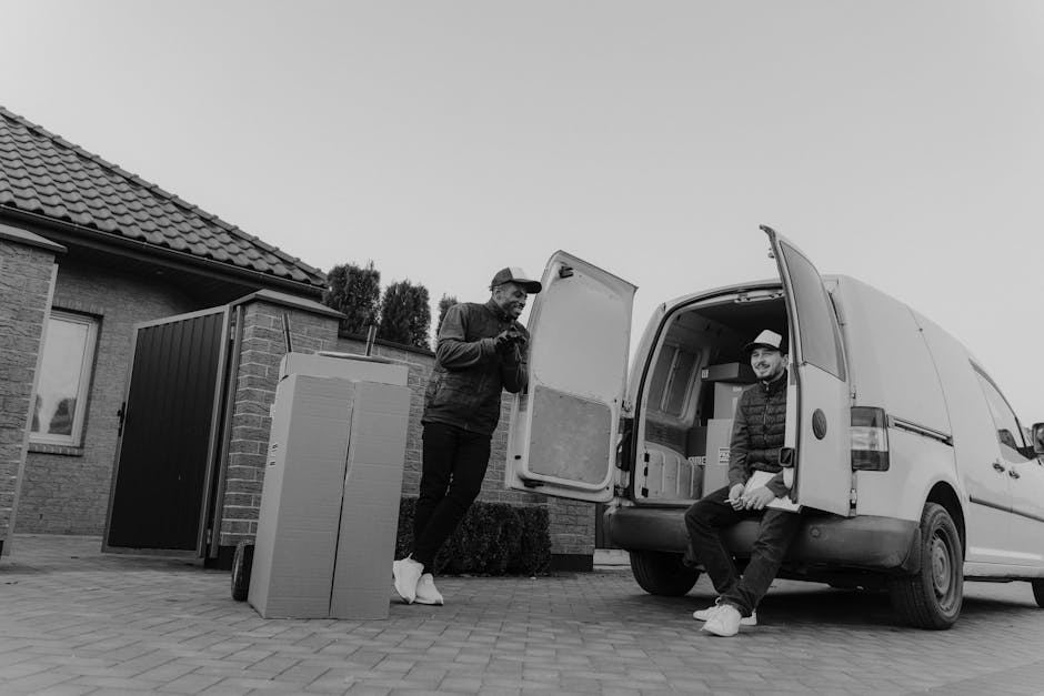Two men are engaged in a home relocation process involving furniture and boxes outside a residential property. One man, standing on the paved driveway, is wearing casual clothing and a cap, and is holding a mobile device while nearby cardboard boxes are positioned on a trolley or dolly. The other man, sitting at the rear of a white work van with its side door open, is dressed in a jacket and a fedora-style hat, and is resting with the van's interior visible behind him, which contains additional packed boxes and moving supplies. The van is parked close to the house, which features brick walls, a tiled roof, and a window, with a small garden area and a gate visible in the background. This scene captures the loading process typical of professional removals by companies such as Man and Van Kennington, illustrating packing, furniture transport, and logistics involved in same-day house moves involving delays and solutions.