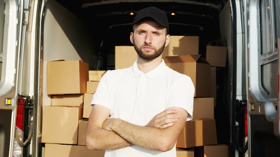 A young man with a beard, wearing a red cap, a beige and brown jacket over a checked shirt, and dark jeans, sits on the open back of a white van used for home relocation. Inside the van, there are several cardboard boxes in various sizes, some stacked on top of each other, with clear packing tape visible on their edges. The boxes are placed on the van’s interior floor, which is partly visible along with the vehicle’s rear doors. The man appears to be resting or pausing during the furniture transport and packing process, which is part of a professional removals service by Man and Van Kennington. The scene is well-lit, with natural light highlighting the arrangements of boxes and the man's neutral expression, reflecting the logistical aspect of loading for a house move or furniture transport. The proximity to the open vehicle emphasizes the loading process involved in home relocation services, with the focus on packing materials, the carrying of goods, and the logistical planning typical of removals undertaken by [COMPANY_NAME].