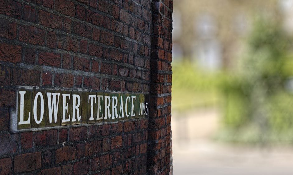 A close-up view of a brick wall with a street sign that reads 'LOWER TERRACE NW3' mounted on it. The sign is composed of individual ceramic tiles with white lettering on a green background. To the right of the sign, the brick wall extends into the background, partially in shadow. In the blurred background, there is greenery and part of a pavement, indicating an outdoor setting. The image appears to be taken during daylight hours, with natural light illuminating the brick and signage. This setting is associated with residential or urban environments typical of house removals and moving services, such as those provided by Man and Van Kennington, highlighting the context of home relocation and furniture transport in the SE11 area for narrow access or terraces.