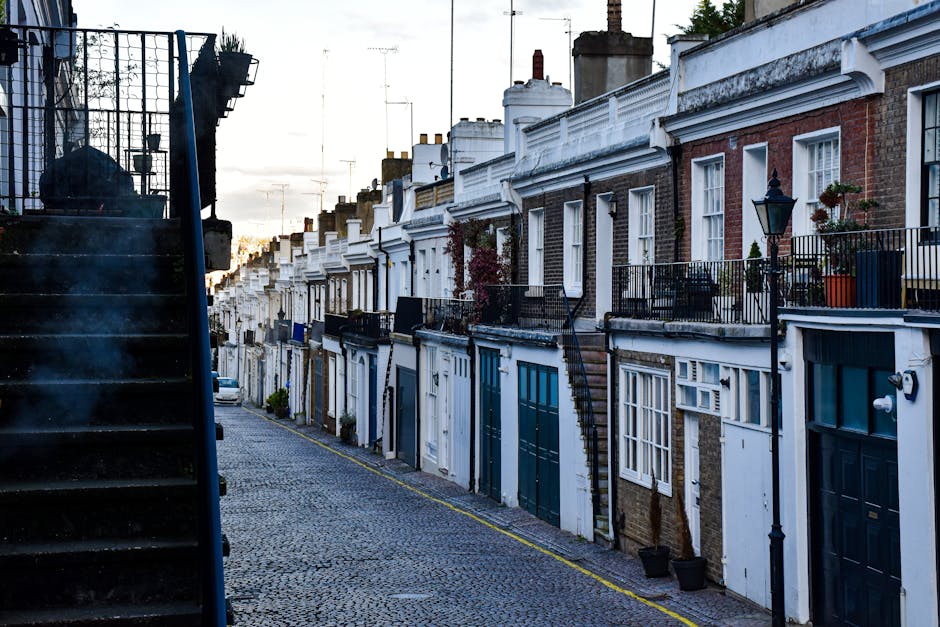 A narrow residential street with a cobblestone pavement in Kennington, featuring terraced brick houses with white and dark-colored painted doors and windows. Each house has a small balcony or front yard enclosed by black metal railings, some adorned with potted plants. On the left side, there is a black metal staircase leading up to a building entrance, partially visible with steps and railing, and smoke rising from a chimney in the background. The scene is captured during the daytime with natural light illuminating the scene, emphasizing the architectural details and the compact layout typical of London terraces. The setting reflects an urban environment suitable for house removals and furniture transport, with a focus on the residential façade, outdoor steps, and street environment, consistent with a home relocation context, as handled by Man and Van Kennington.