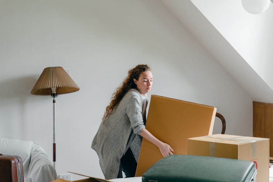 A woman with long, curly hair, wearing a grey cardigan, is inside a room preparing for a home relocation. She is holding a large cardboard box, which is one of several packed boxes placed on a table or surface nearby. The room has a minimalist decor with white walls, a wooden lamp with a beige pleated fabric shade, and a piece of furniture partially visible on the left. The space appears to be an attic or upper-floor room, suggested by the sloped ceiling and skylight window. The woman is in the process of packing or unpacking, and her expression is focused. Behind her, the environment suggests a moving or packing activity, possibly linked to furniture transport or packing and moving services. The scene reflects the preparatory steps involved in a house removal, with items carefully boxed in preparation for transportation by a relocation service such as Man and Van Kennington, which provides domestic removals and delivery solutions.