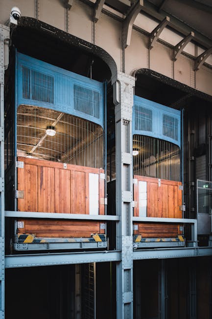 Two wooden-framed elevator cabins with metal mesh upper panels are visible inside a building, designed for freight and passenger transport. The cabins are positioned on upper levels, with a vertical metal framework supporting them, and appear to be part of a home relocation or moving logistics setup. The surrounding environment includes a high ceiling with exposed metal beams, and the lighting is industrial. The image emphasizes the loading process, with no visible people or moving equipment, but the spacious interior suggests preparation for furniture or large items to be moved between floors, in line with professional removals services like those offered by Man and Van Kennington. This setting illustrates an alternative to traditional stairs for moving household goods, supporting efficient internal transport during a house move or relocation, especially when lifts are unavailable or fail, aligning with the service details in the page titled 'When lifts fail Kennington removals for upper floor homes.'